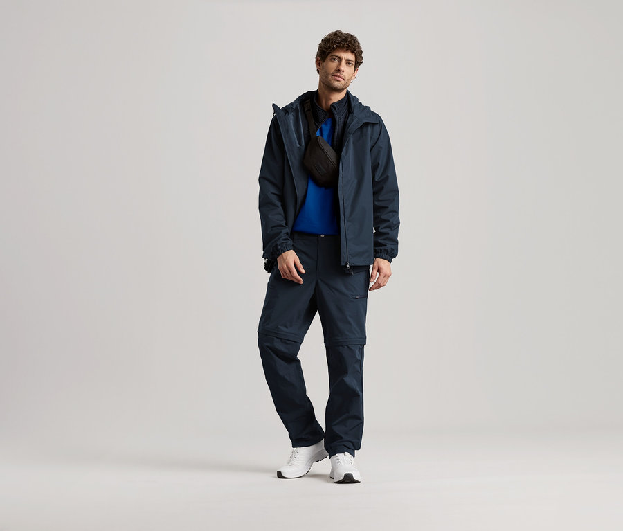 Egy férfi áll sötétkék kabátban, nadrágban és fehér tornacipőben, fekete oldaltáskával.