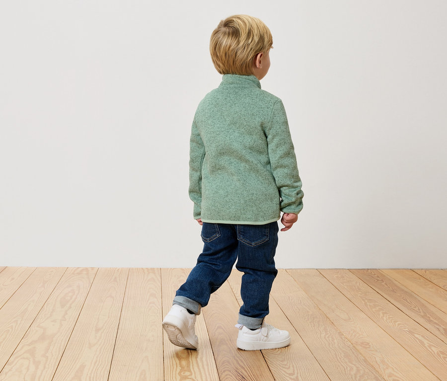 Egy gyermek háttal a kamerának sétál egy zöld polár pulóverben, farmerban és fehér tornacipőben egy fapadlón.