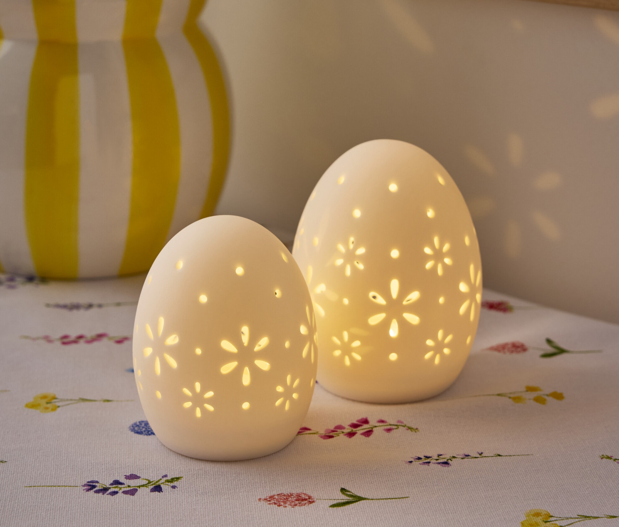 Két fehér, tojás alakú, virágmintás lámpa áll egy asztalon, a háttérben egy virágváza látható.