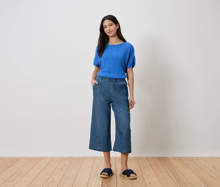 Nő áll kézzel a zsebében, kék felsőt, farmer culottes-t és szandált visel egy fehér fal előtt.