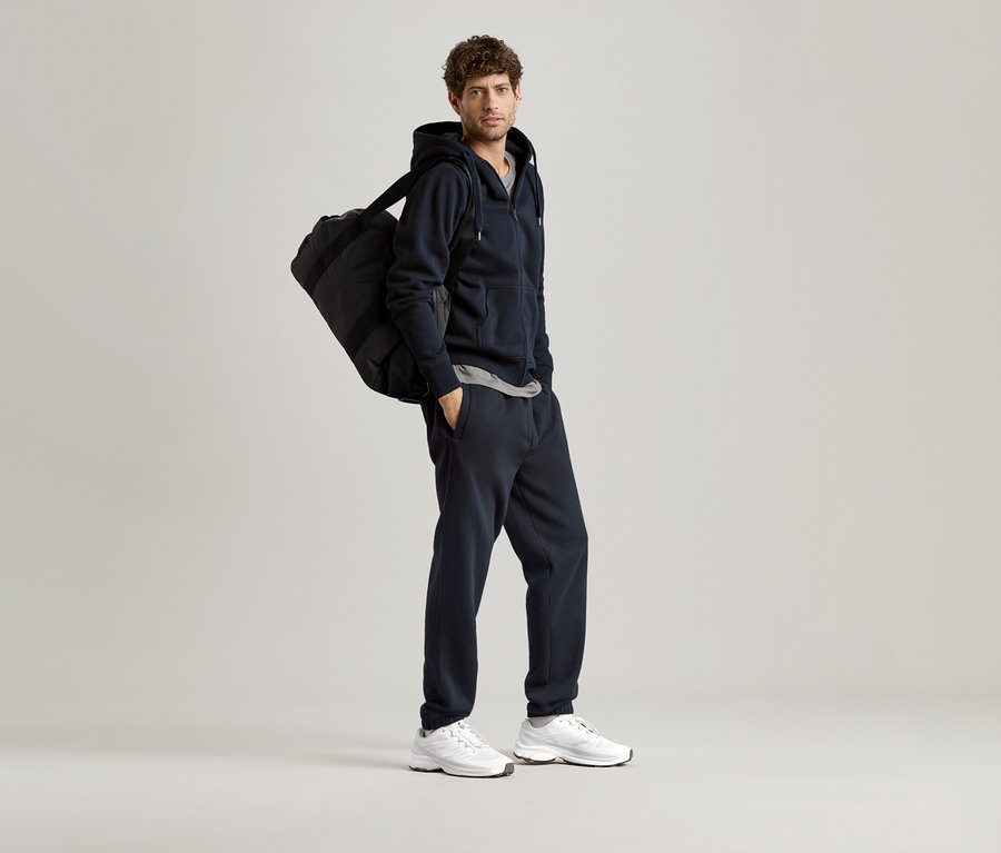 Férfi sötétkék kapucnis pulóverben, sporttáskával a vállán.