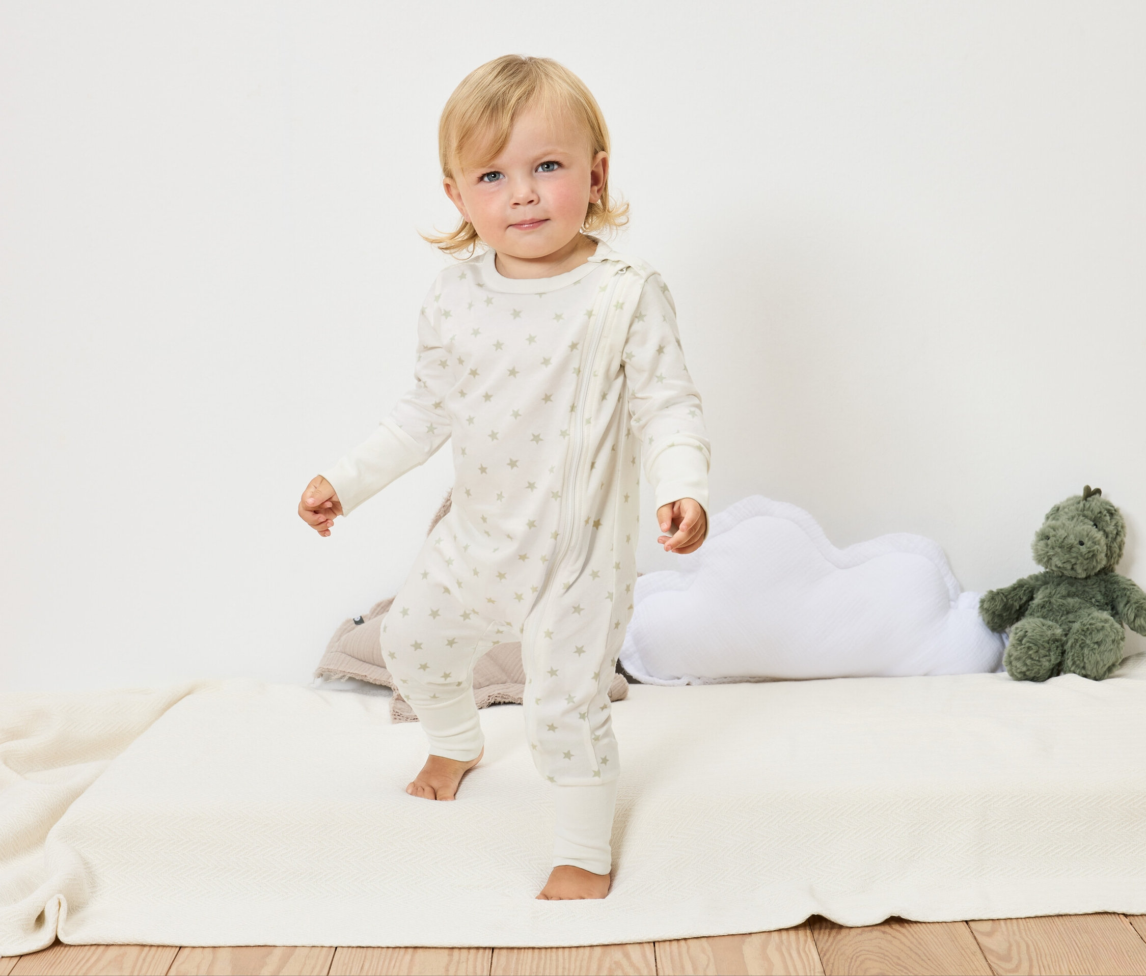 Egy szőke kisgyermek csillagos, fehér overallban áll mezítláb egy fehér takarón egy felhőpárna és egy zöld plüssállat mellett.