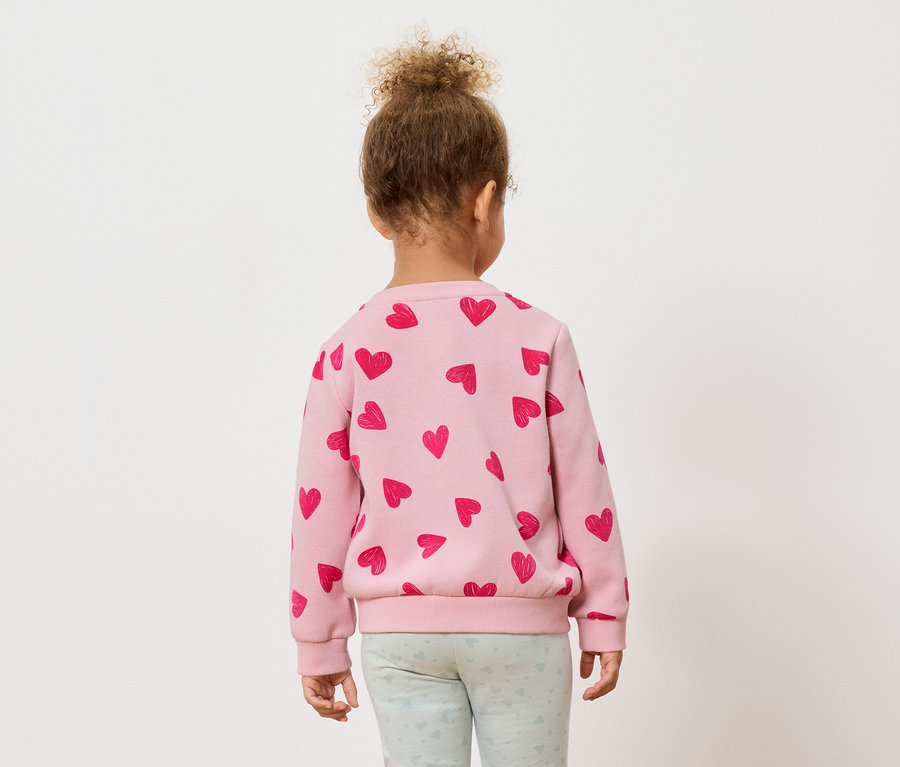 Hátulnézet egy kisgyermekről, aki göndör hajjal rendelkezik, rózsaszín pulóvert visel, piros szívekkel, és világoskék leggings-et.