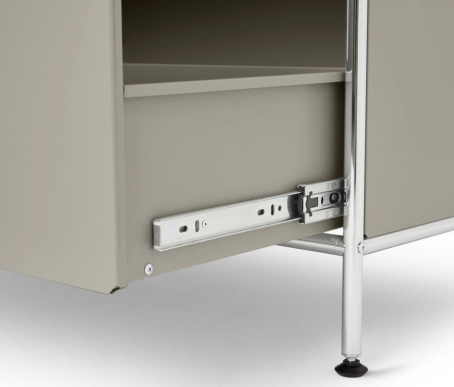 Egy szürke szekrény közeli képe részben kihúzott fiókkal, ezüstszínű fém sínekkel és egy krómozott fém lábbal.