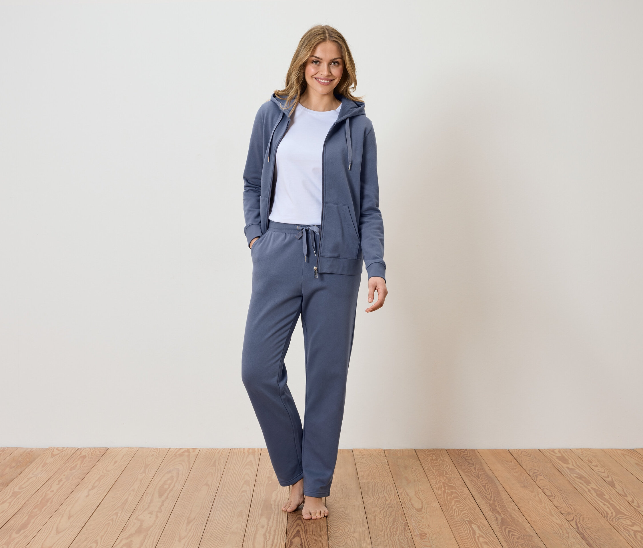 Nő áll mezítláb a fapadlón, kék tréningruhát visel, kapucnis pulóverrel és fehér pólóval.