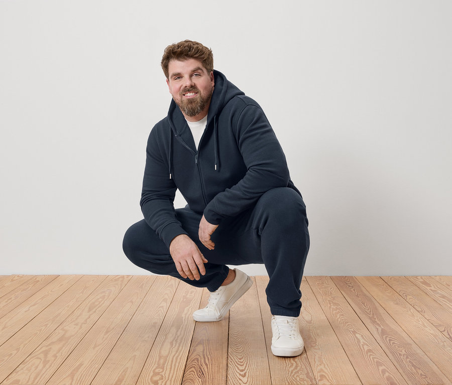 Férfi guggol a fapadlón, sötétkék tréningruhát és fehér tornacipőt visel.