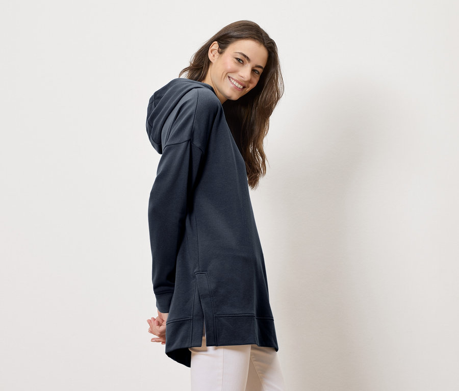 Nő hosszú, tengerészkék kapucnis pulóvert és világos nadrágot visel, a kamerába mosolyog.