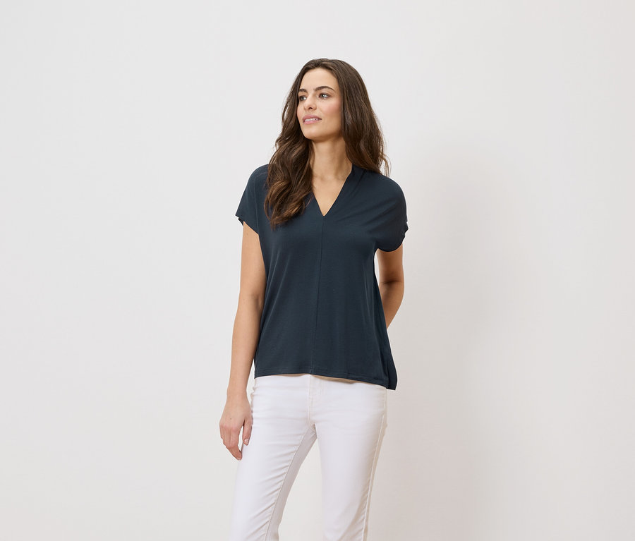 Nő sötétkék pólót és fehér nadrágot visel fehér háttér előtt.