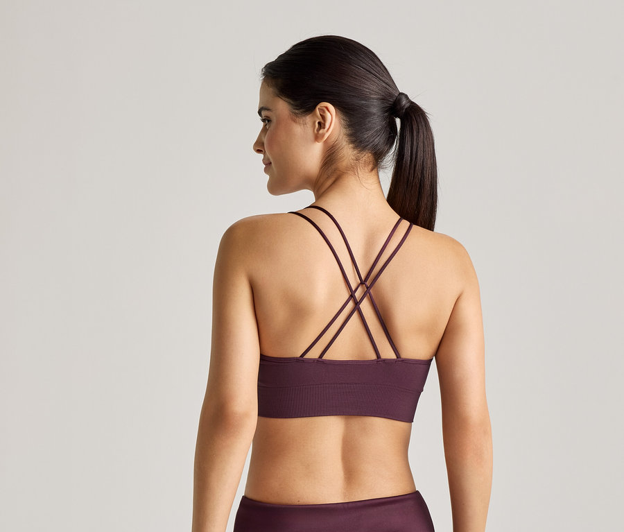 Hátulról látható nő, lila, keresztezett pántos sportmelltartót és leggingset visel világos háttér előtt.