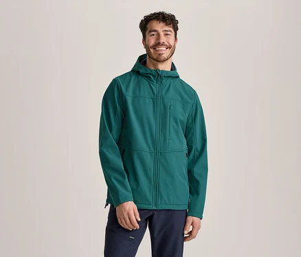Egy mosolygós, göndör hajú és szakállas férfi áll szemből, zöld kapucnis softshell kabátot és sötétkék nadrágot visel.
