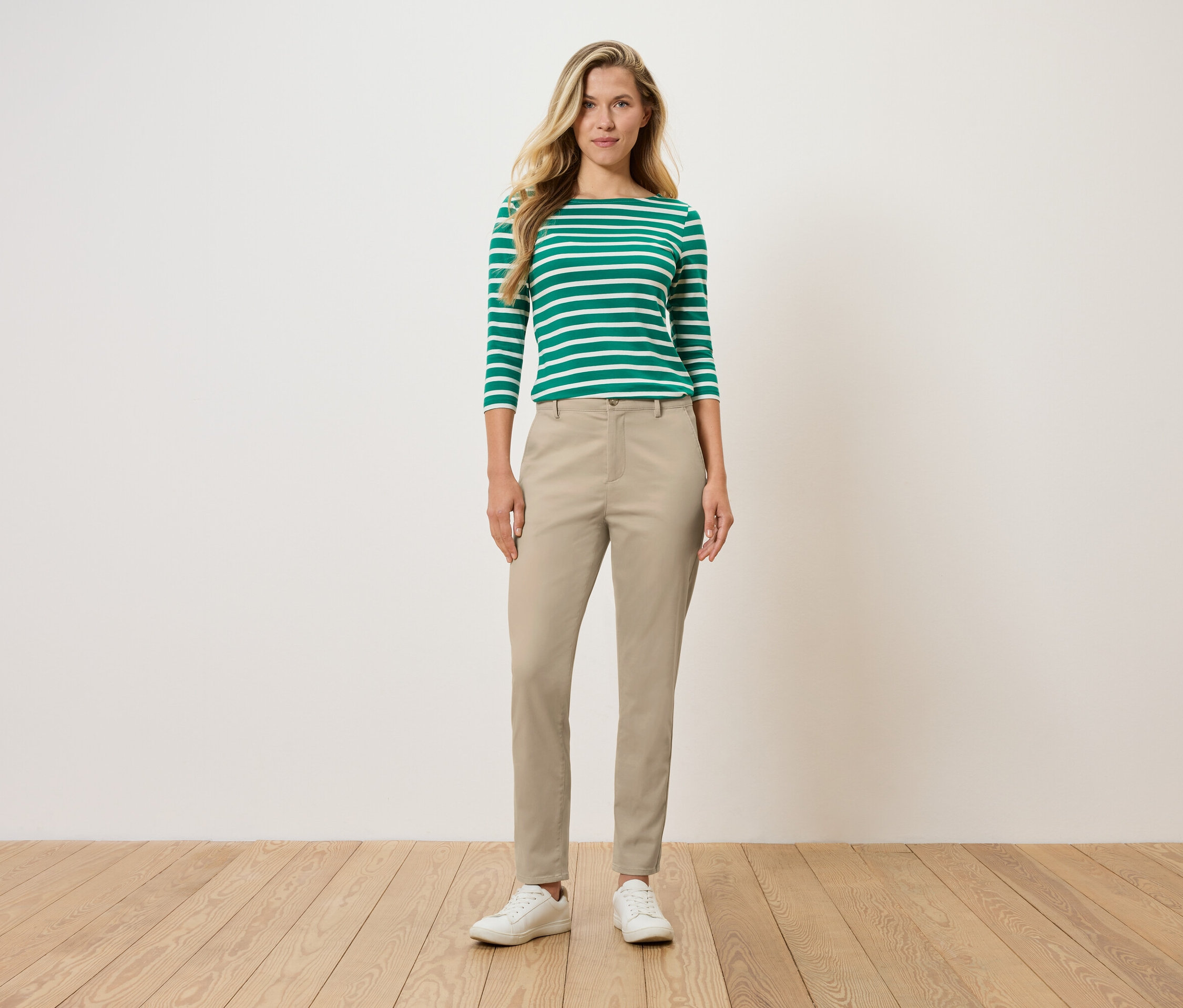 Egy nő áll, zöld-fehér csíkos hosszú ujjú inget, bézs nadrágot és fehér cipőt visel.