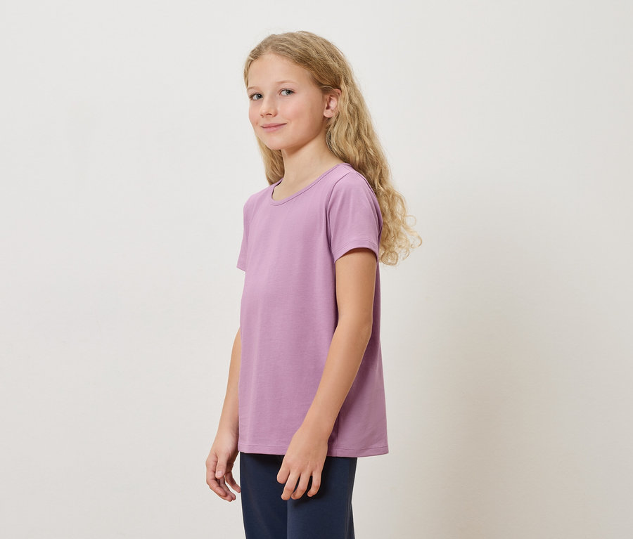 Egy hosszú szőke hajú lány lila pólóban és sötétkék nadrágban áll fehér háttér előtt.