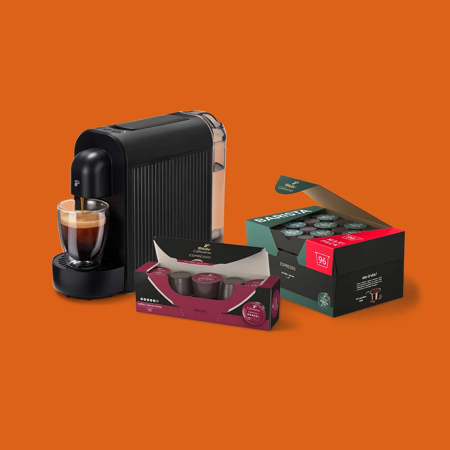 Fekete kávéfőző önt espressót egy üvegcsészébe, mellette egy nyitott doboz 96 Barista Espresso kapszulával és egy kisebb doboz Tchibo Cafissimo Espresso kapszulával.