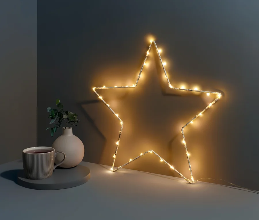 Egy kis LED-es fémcsillag áll egy asztalon egy csésze és egy váza mellett.