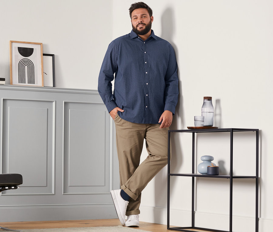 Egy férfi farmer hatású Comfort Fit ingben, barna chino nadrágban és sötétkék merinó zokniban (4 pár, sötétkék, piros és sötétzöld) áll egy szobában.