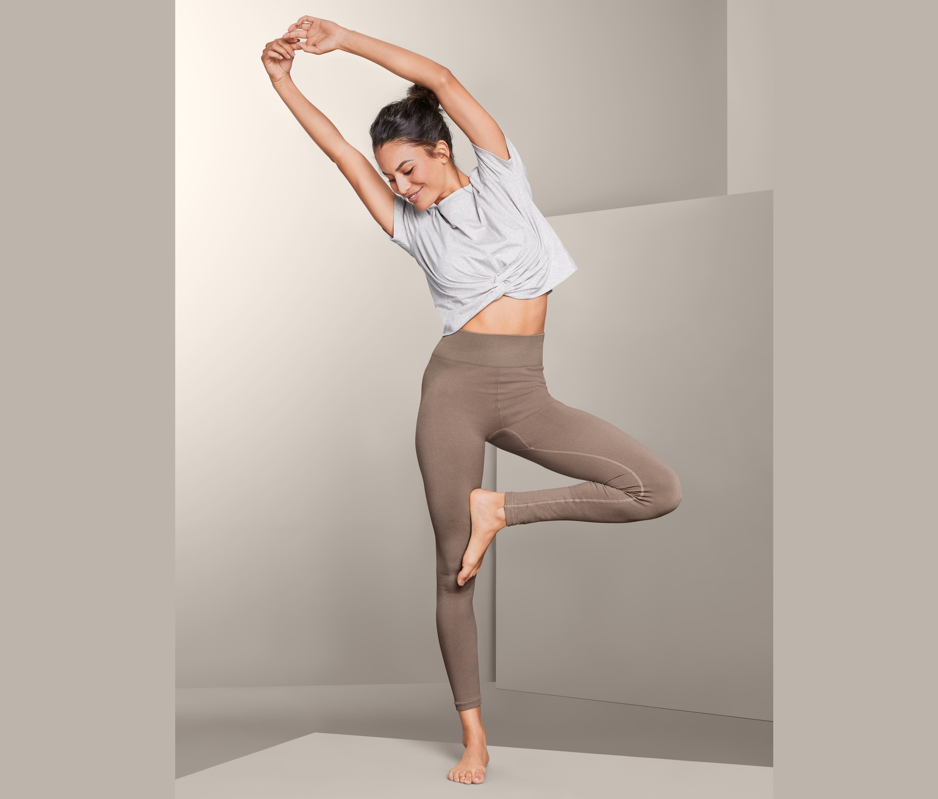 Nő világosbarna, alakformáló, seamless sportnadrágban egy lábon áll, karjai a feje fölött.