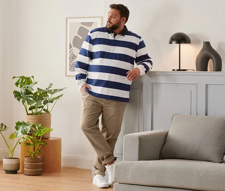 Férfi támaszkodik a falnak barna chino nadrágban, sötétkék/fehér csíkos hosszú ujjú pólóingben és fehér sneaker cipőben.