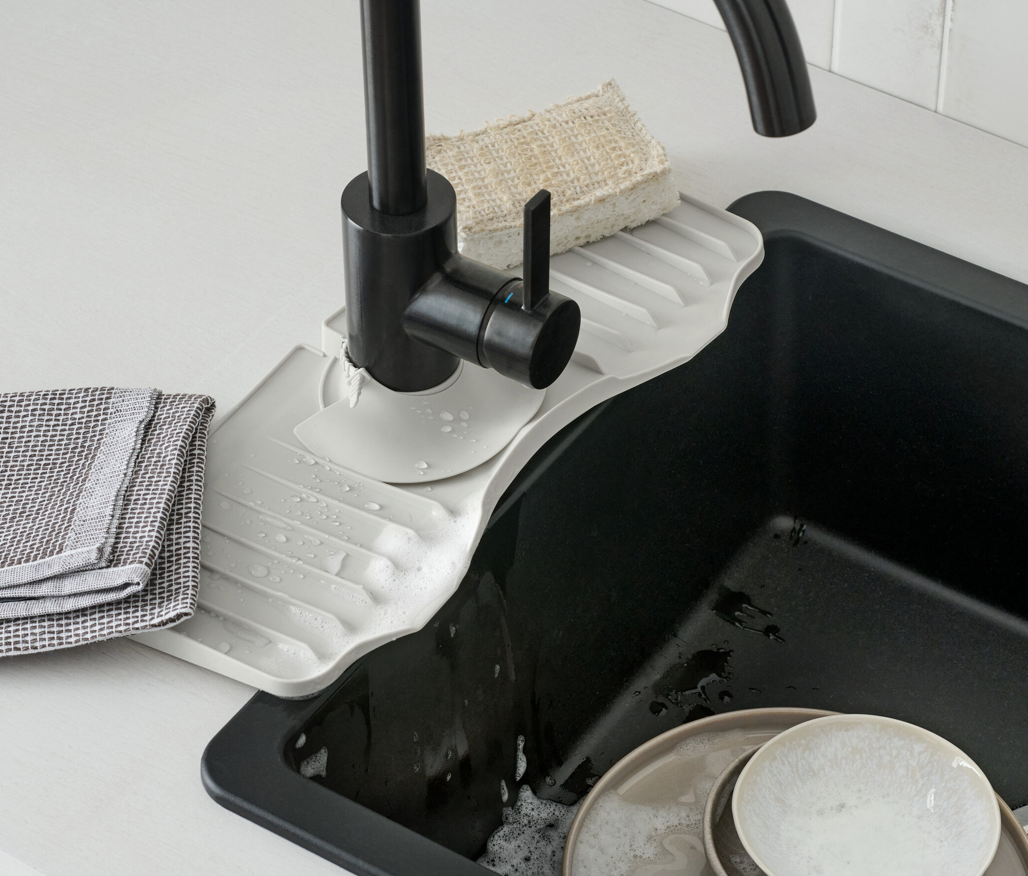 Szilikon cseppfogó vízcsaphoz található egy fekete csap alatt. Piszkos edények vannak a mosogatóban.