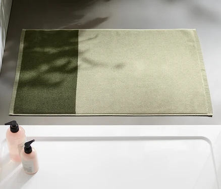 Egy szürkészöld-mohazöld jacquard fürdőszobaszőnyeg látható a fürdőszobában, a zuhanyzó mellett.