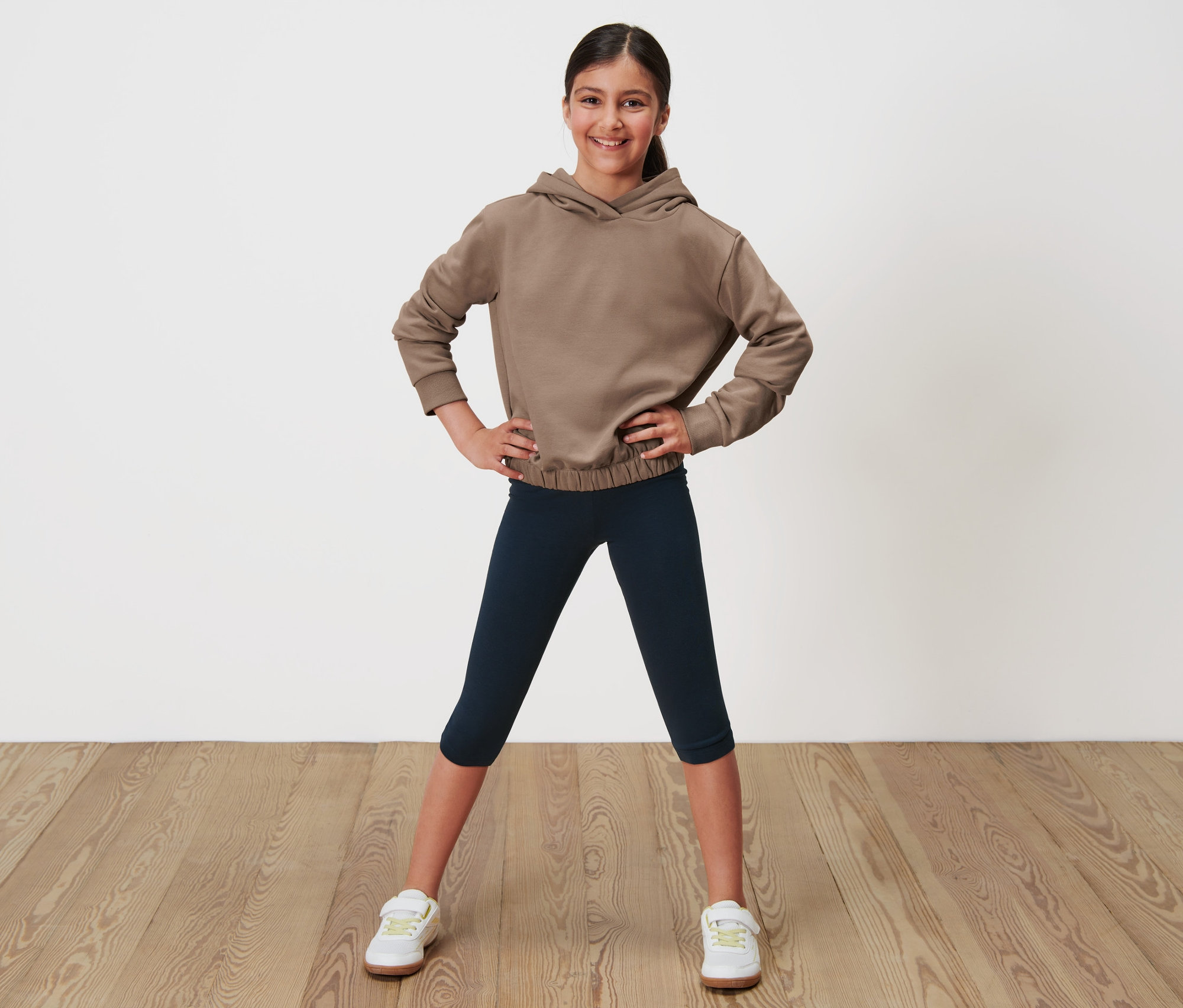 Lány pózol barna kapucnis pulóverben és sötétkék 3/4-es leggingsben.