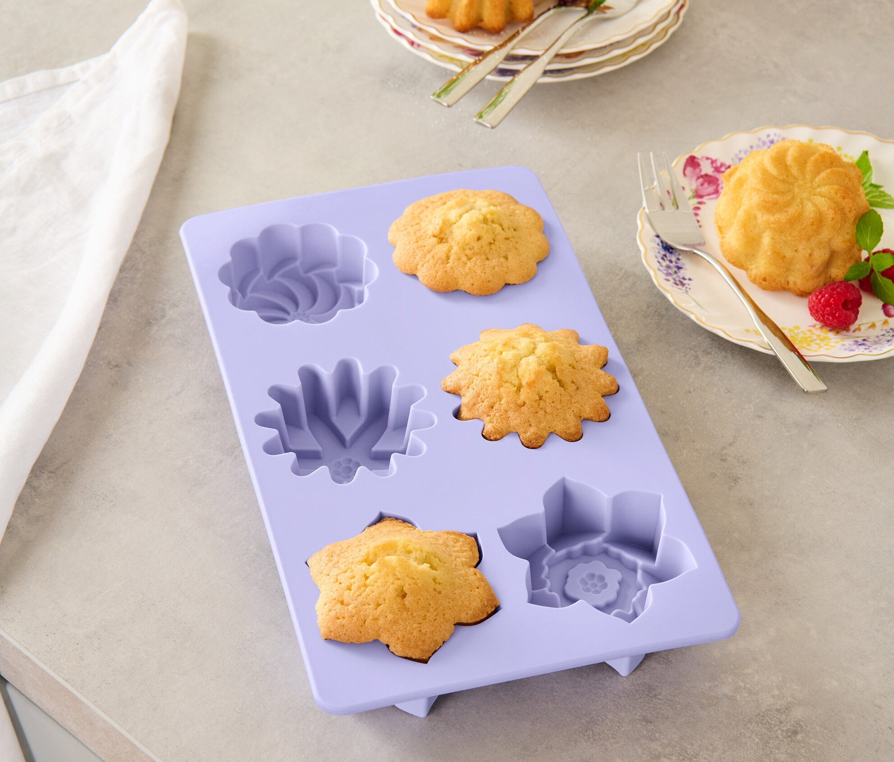 Lila virág alakú sütőforma sült muffinokkal, két muffin tányérokon villákkal.