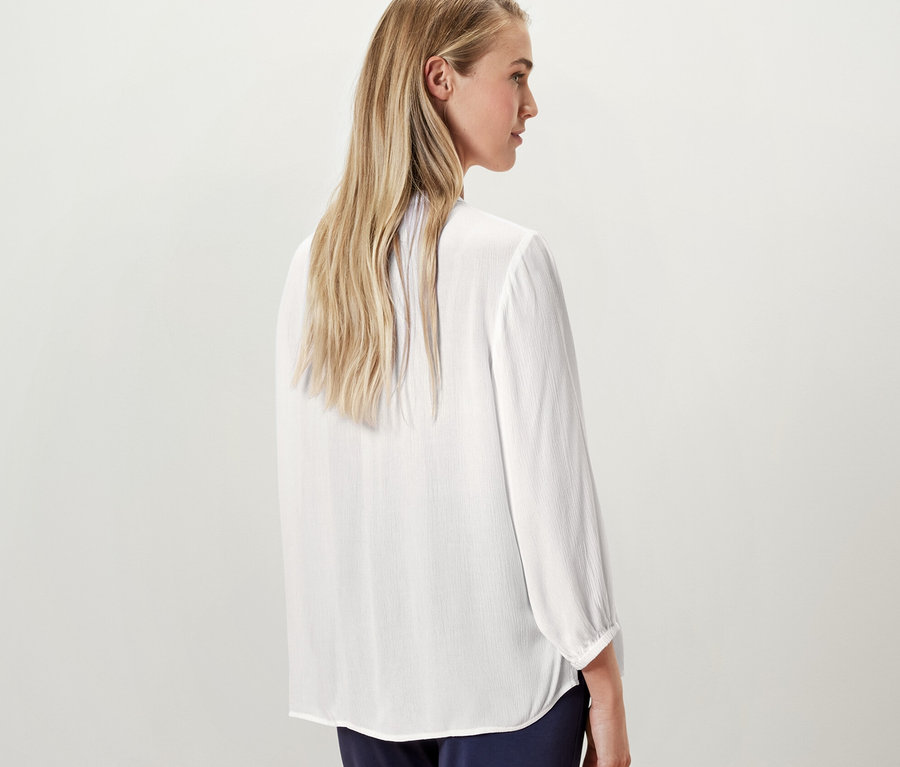 Egy szőke nő a kamerának háttal állva pózol, és egy fehér női 7/8-os ujjú tunikablúzt visel.