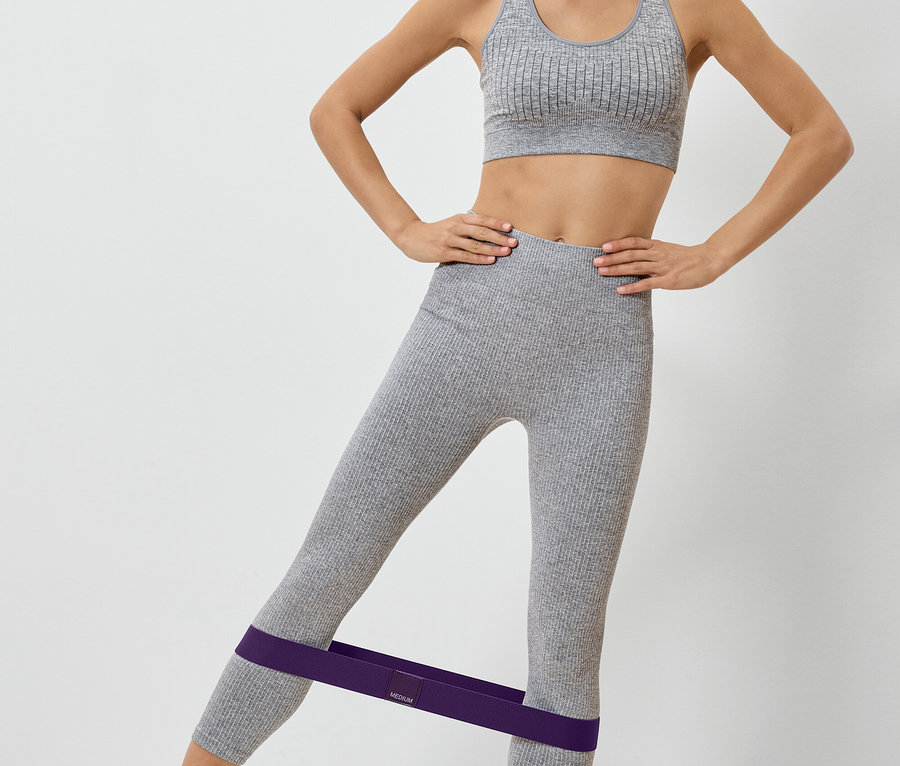 Nő szürke sportruhában, lila gumiszalaggal a lábán edz.