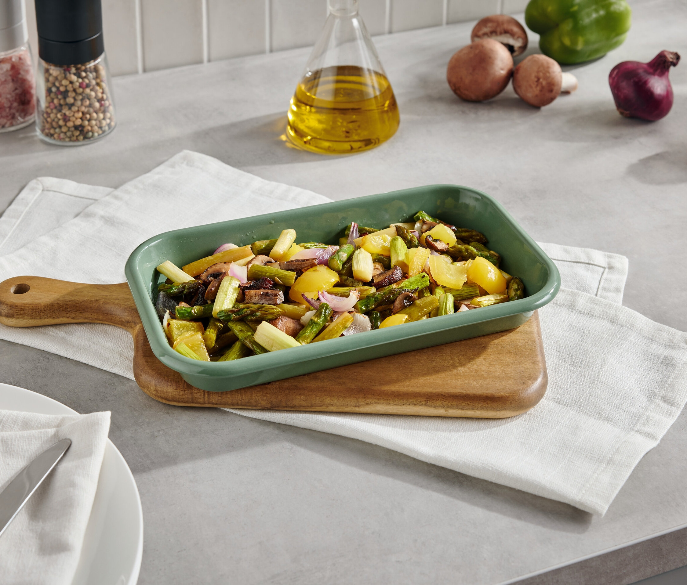 Zöld rakott edényben sült zöldségek deszkán szalvétával. A háttérben gombák, paprika, hagyma és fűszerek találhatók.