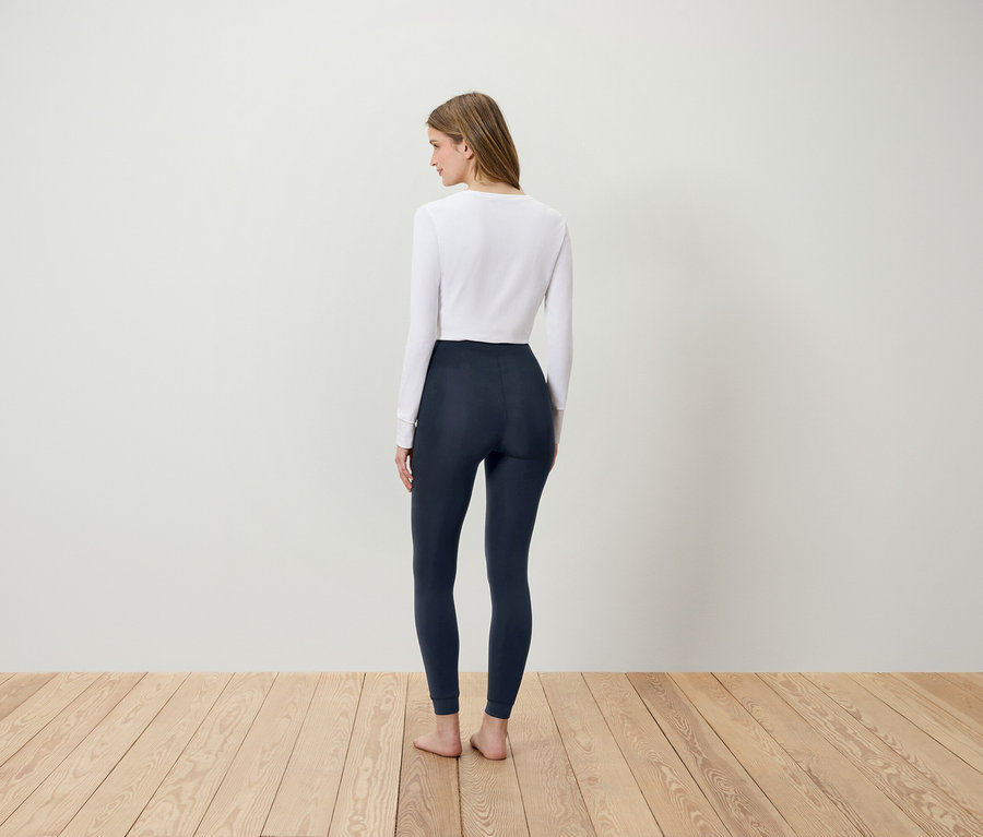Nő áll fehér felsőben és sötétkék leggingsben, háttal a nézőnek, fapadlón.