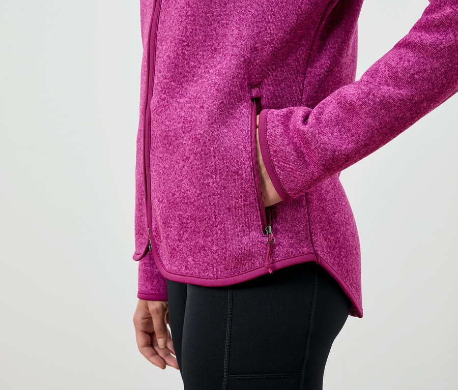 Nő a kezét egy rózsaszín polárkabát zsebében tartja, fekete leggingset visel.