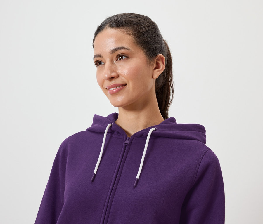 Barna hajú, lófarokba kötött nő lila, cipzáras kapucnis pulóvert visel.