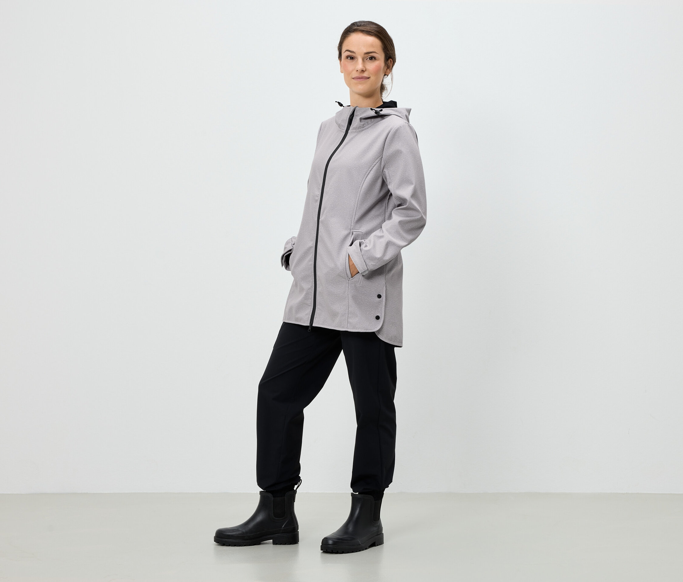 Egy nő áll, kezét egy szürke kapucnis kabát zsebében tartja, fekete nadrágot és fekete csizmát visel.