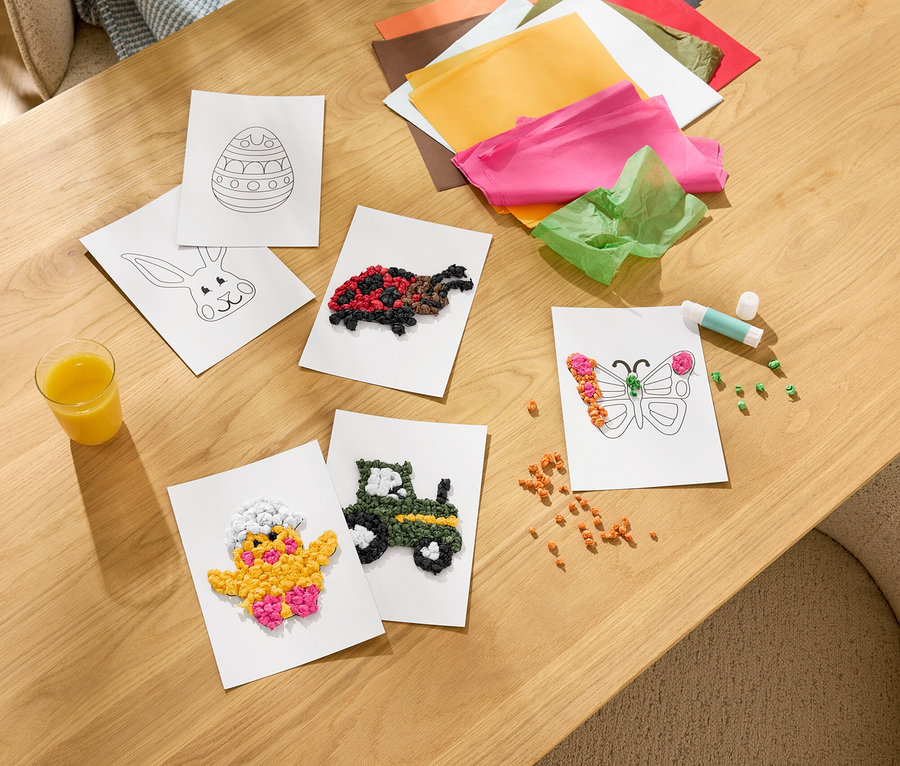 Egy fa asztalon kézműves alkotások találhatók, köztük ragasztott csibe-, traktor-, pillangó- és teknősbéka kártyák. Színes selyempapír, ragasztórúd és egy pohár gyümölcslé is látható.