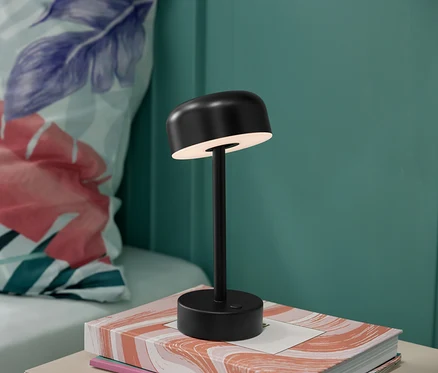 Egy fekete LED-es akkus asztali lámpa egy könyvön áll, a háttérben virágos párna látható.