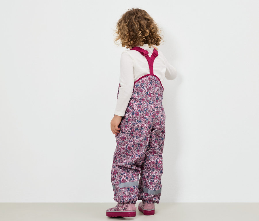 Virágos kertésznadrágot és rózsaszín csizmát viselő lány háttal áll a kamerának.