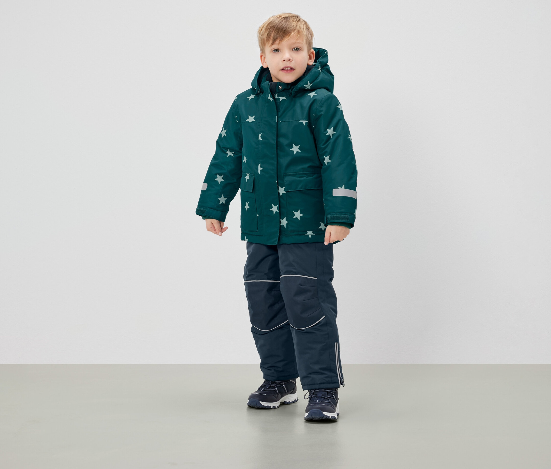 Fiú áll csillagos, zöld téli kabátban és sötétkék nadrágban.