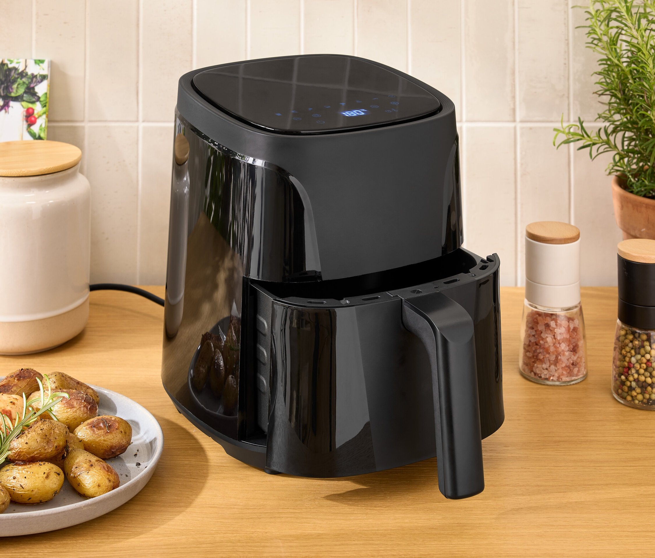 Egy fekete forrólevegős fritőz áll egy fa asztalon egy tányér csirke és burgonya mellett.