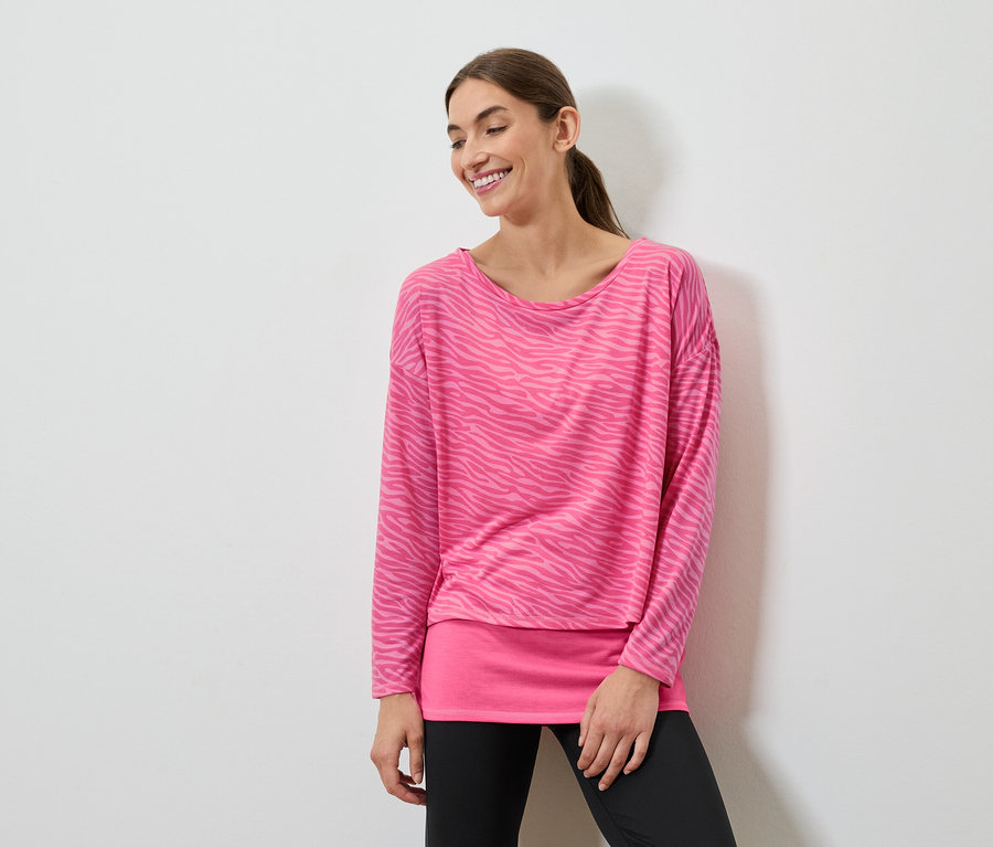Mosolygó nő pózol rózsaszín, hosszú ujjú felsőben és fekete leggingsben.