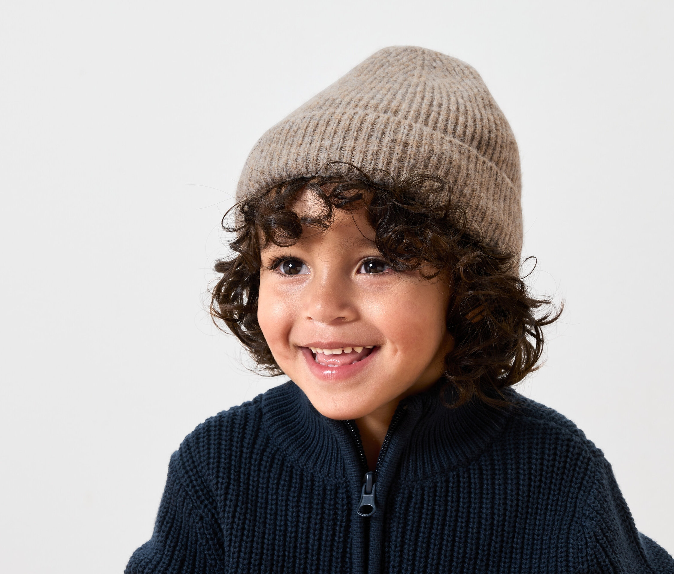 Mosolygó, barna, göndör hajú fiú bézs színű gyapjú sapkát és sötétkék, kötött, cipzáras kardigánt visel.