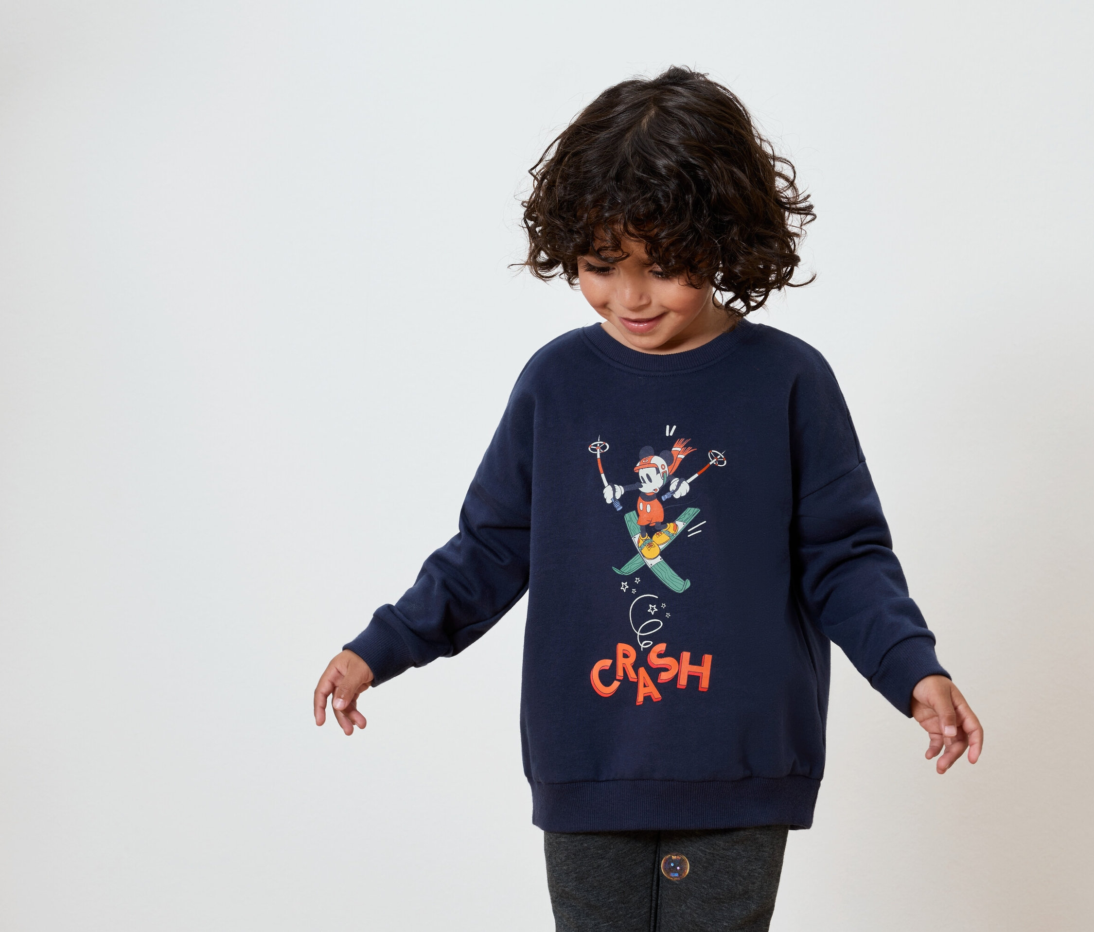 Egy sötét, göndör hajú gyermek sötétkék pulóvert visel, amelyen Mickey egér síelés közben látható.