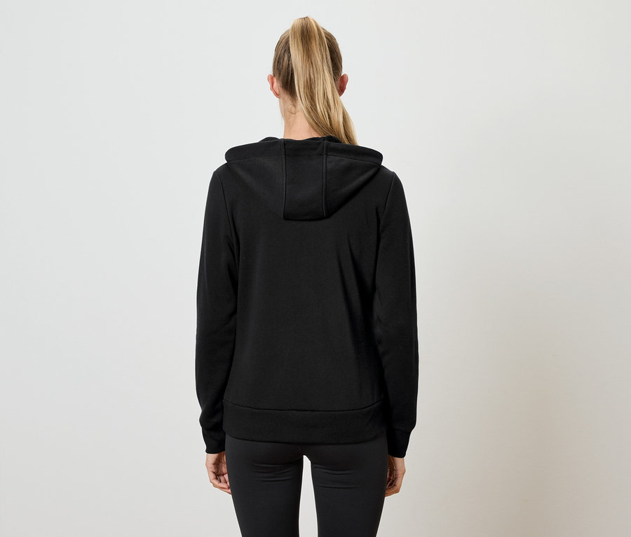 Hátulnézetből egy szőke hajú, lófarokba kötött nő látható, fekete kapucnis pulóvert és fekete nadrágot visel.