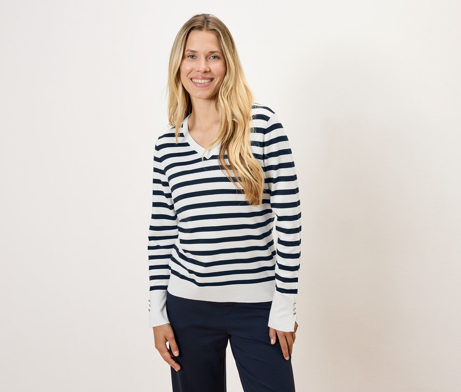 Egy hosszú szőke hajú nő csíkos pulóvert és sötétkék nadrágot visel.