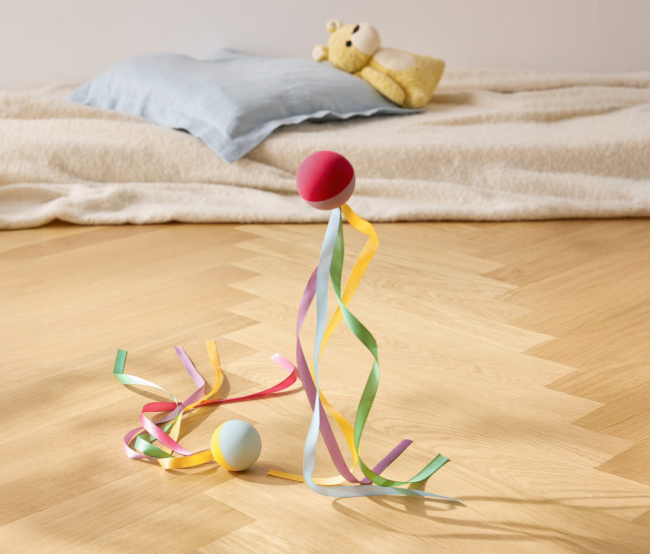 Egy piros labda színes szalagokkal áll egy fapadlón, a háttérben egy párna és egy plüssmackó.