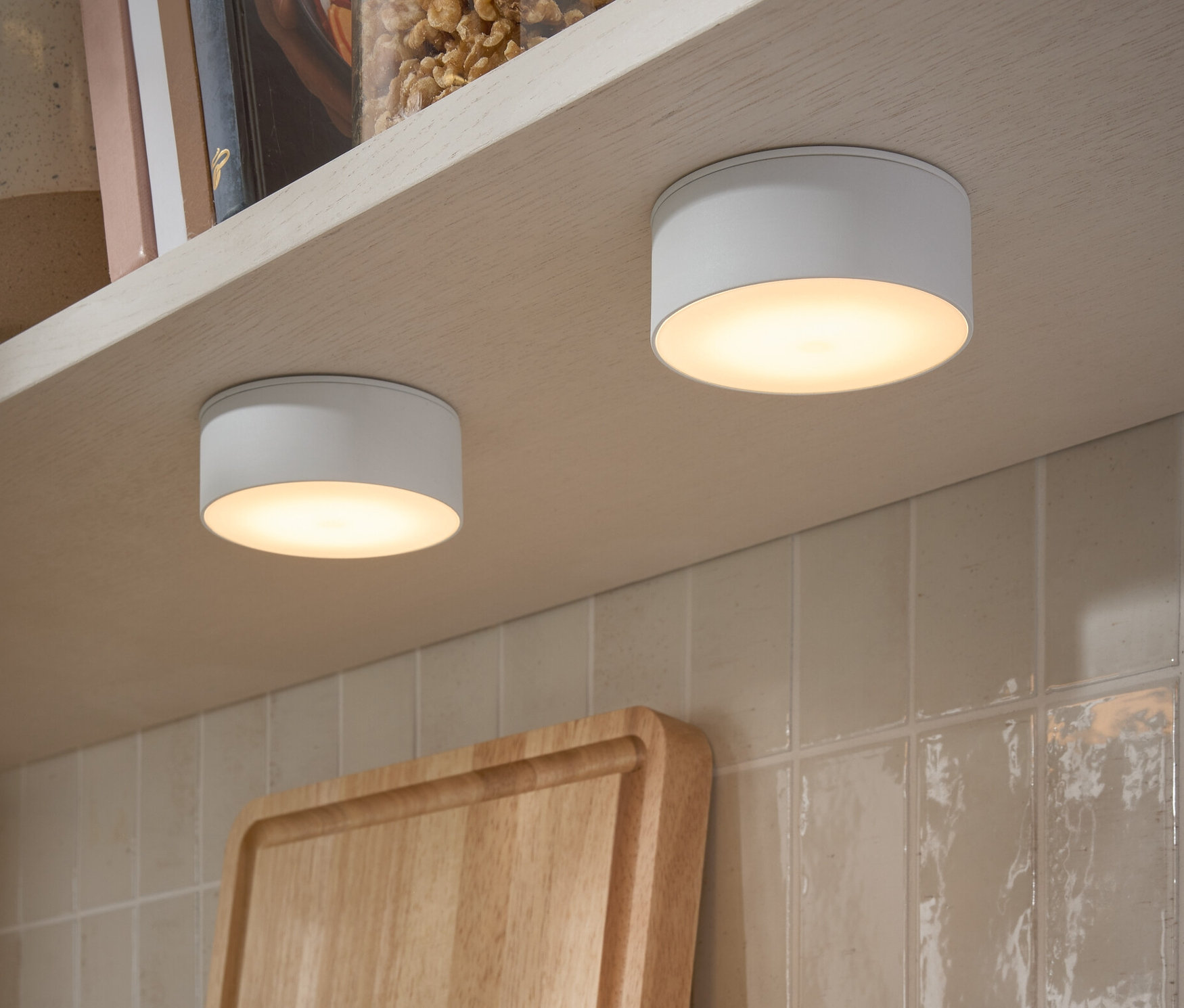 Két LED-es multifunkciós lámpa van rögzítve egy polc aljára. A polcon diós üvegek és könyvek találhatók.