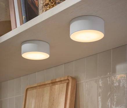 Két LED-es multifunkciós lámpa van rögzítve egy polc aljára. A polcon diós üvegek és könyvek találhatók.