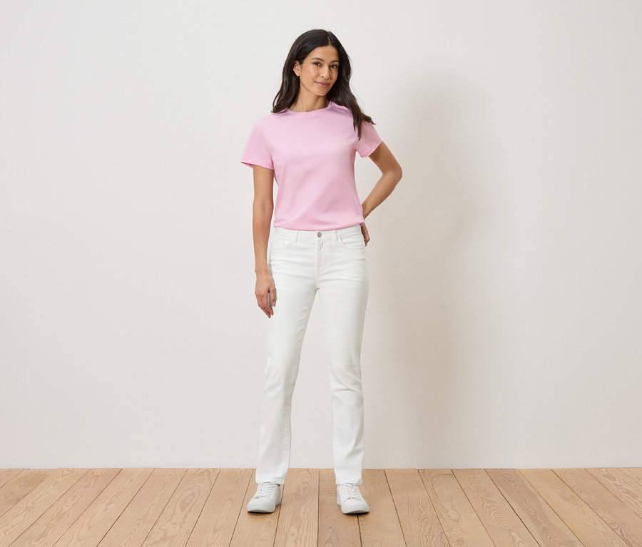 Nő áll egy fehér fal előtt, rózsaszín pólót, fehér farmert és fehér tornacipőt visel.