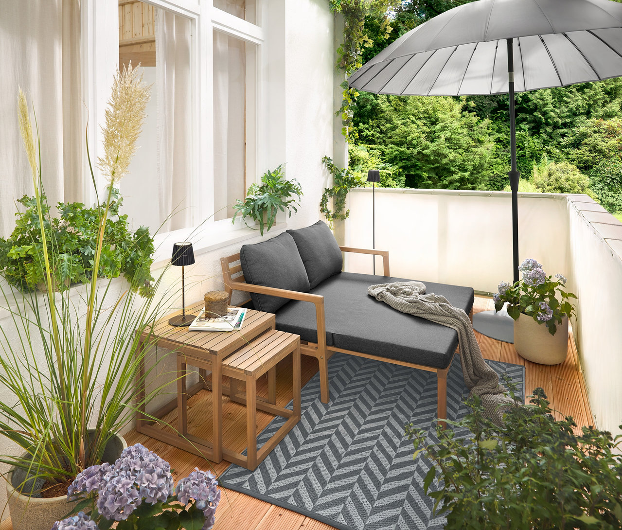 Elrendezés egy erkélyen kihúzható kerti kanapéval, kültéri szőnyeggel (kb. 165 x 115 cm), krémszínű belátásgátló és szélvédő erkélyre, fekete LED-es akkumulátoros kültéri asztali lámpával és fekete LED-es akkumulátoros kültéri állólámpával.