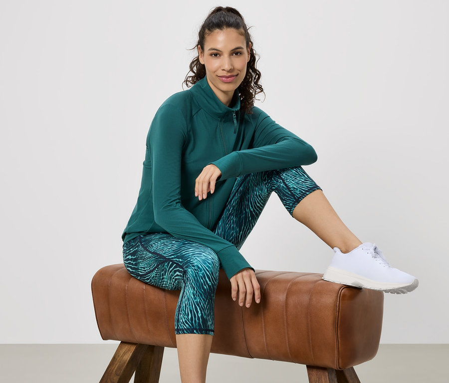 Nő ül egy tornabakon, zöld sportkabátot, mintás leggingset és fehér sportcipőt visel.