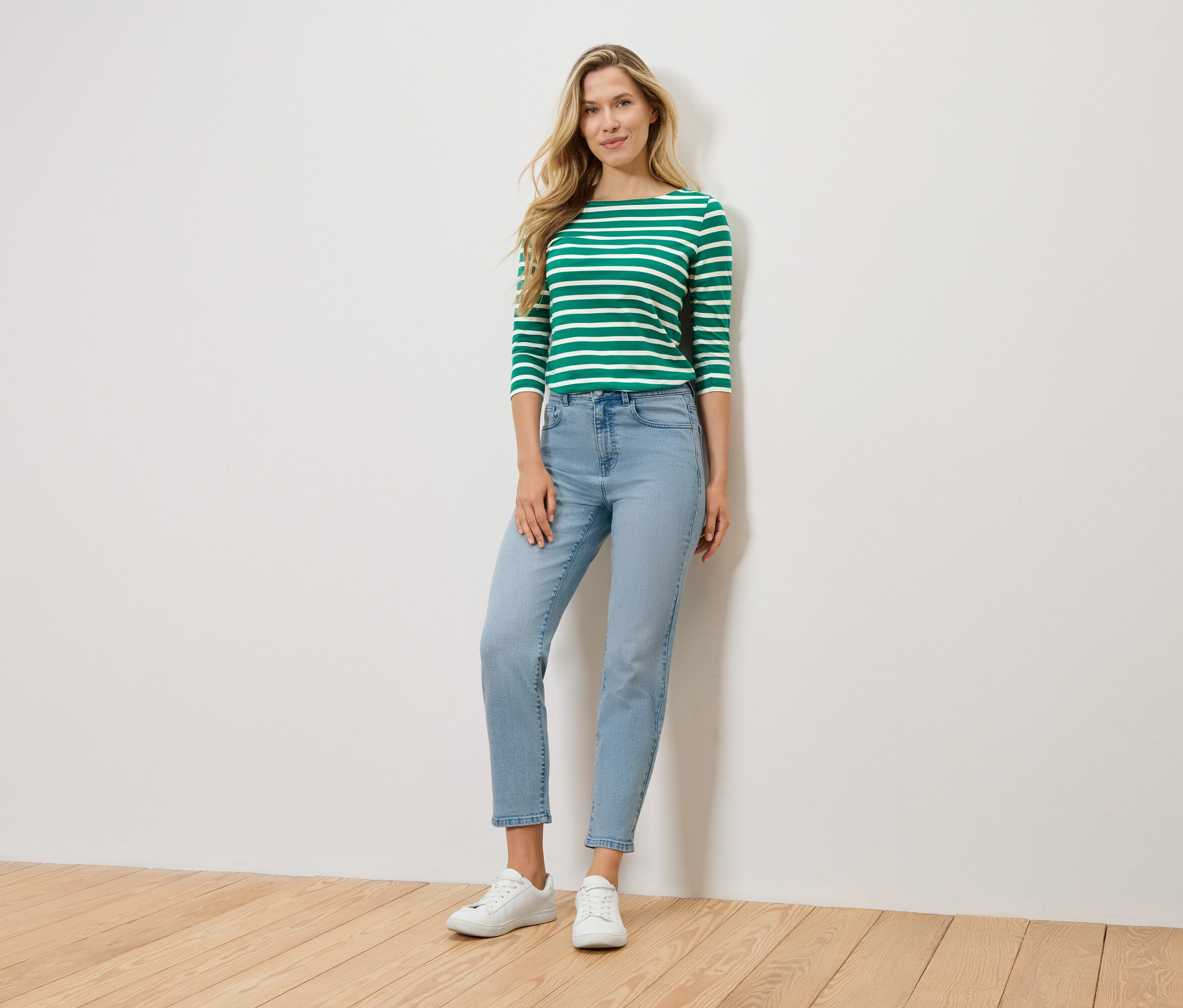 Egy szőke nő áll egy fal előtt, zöld-fehér csíkos, hosszú ujjú felsőt, világoskék farmert és fehér tornacipőt visel.