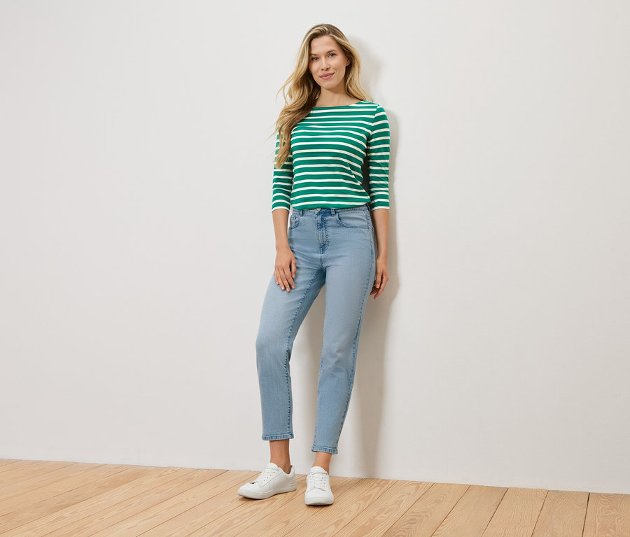 Egy szőke nő áll egy fal előtt, zöld-fehér csíkos, hosszú ujjú felsőt, világoskék farmert és fehér tornacipőt visel.